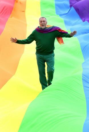 Gilbert Baker walking on a giant rainbow flag.